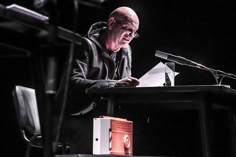 Andreas Keller sitzt bei einer Lesung an einem Tisch. Im Vordergrund steht ein historisches Kofferradio.