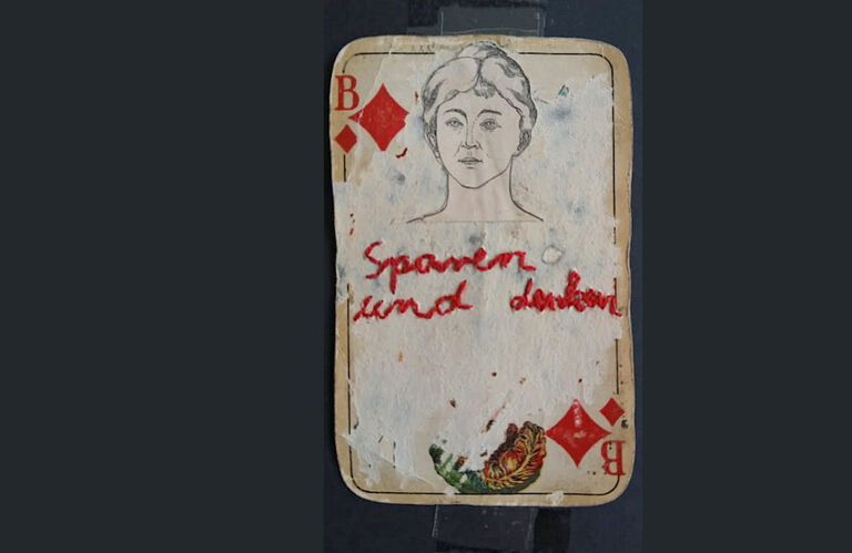 Auf einer Collage aus einer Spielkarte und der Zeichnung eines Gesichtes wurde mit rotem Garn der Schriftzug "Sparen und Denken" gestickt.