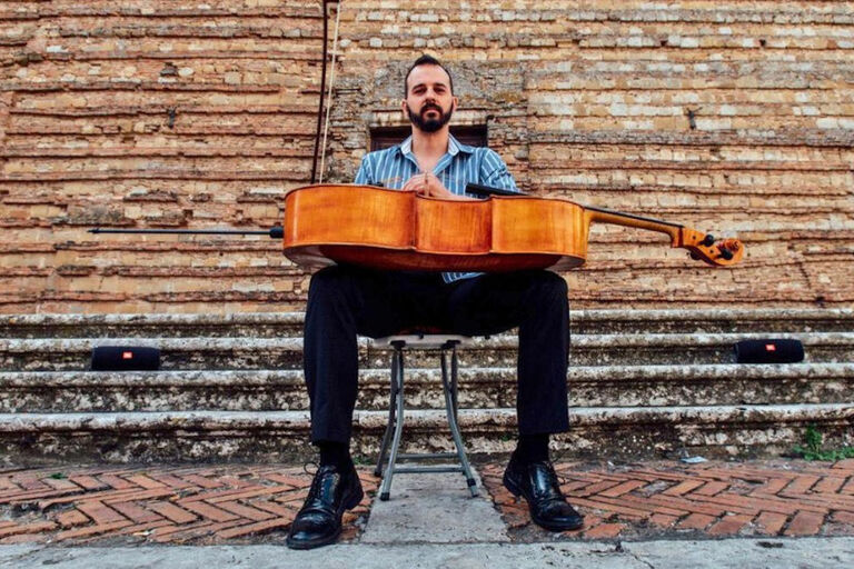 Der Musiker sitzt mit seinem Cello auf den Knien vor einem historischen Gebäude.