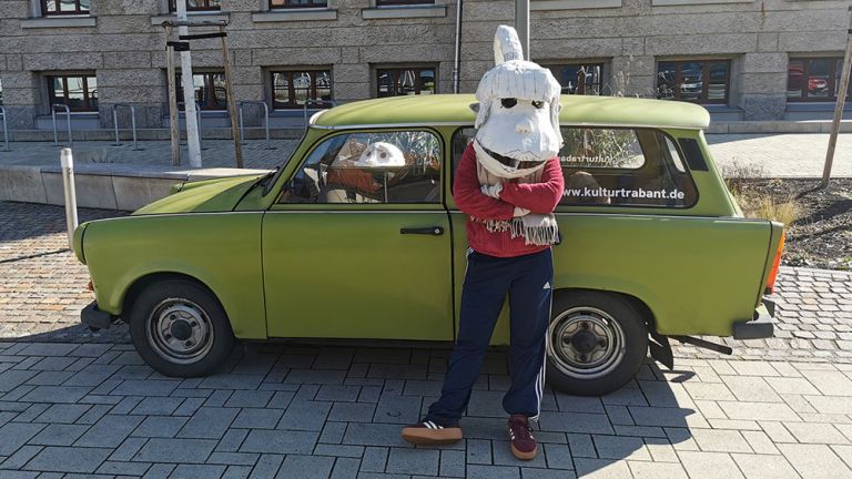 Eine Person steht mit einer überdimensionierten weißen Maske vor einem Trabant