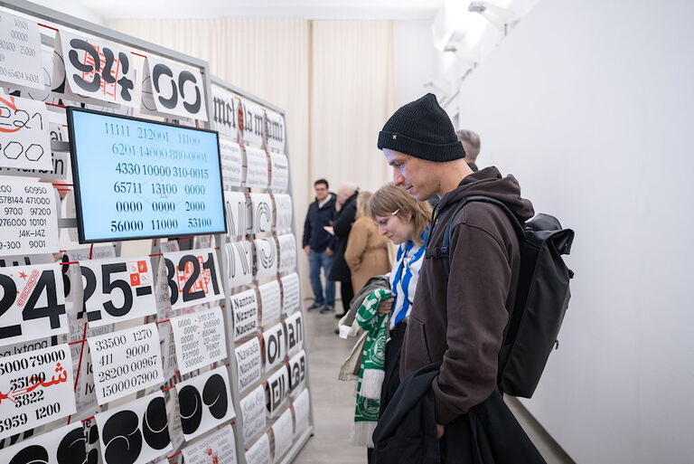 Mehrere Menschen stehen vor Stelwänden mit verschiedenen Buchstaben und Zahlen auf einzelnen Papierblättern.