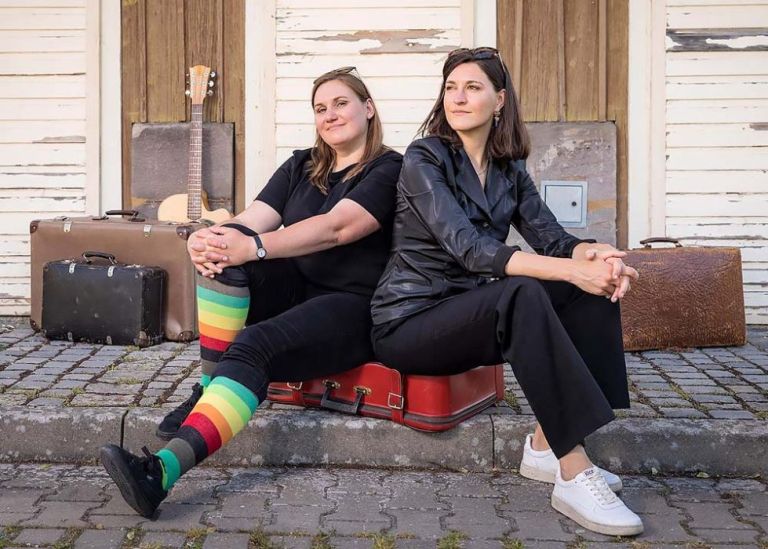 Die beiden Musikerinnen sitzen auf einem Koffer vor einer Holzbaracke mit abblätternder Farbe.