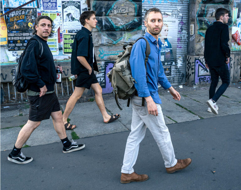 Bandfoto, das wie ein Schnappschuss der Musiker wirkt, die an einer Hauswand mit Plakaten und Graffiti entlanglaufen