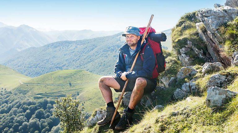Devid Striesow sitzt als Hape Kerkeling in seiner Wanderausrstung auf einem Felsen mit einem weiten Ausblick auf einer Berglandschaft.