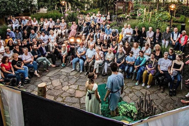 Viele Zuschauerinnen und Zuschauer sitzen im Garten auf Stühlen um eine kleine Bühne.