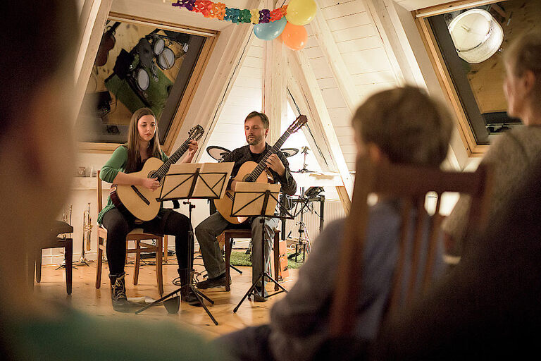 Eine Frau und ein Mann sitzen in einem Dachzimmer und spielen für Publikum, das auf Sthlen sitzt, Gitarre.