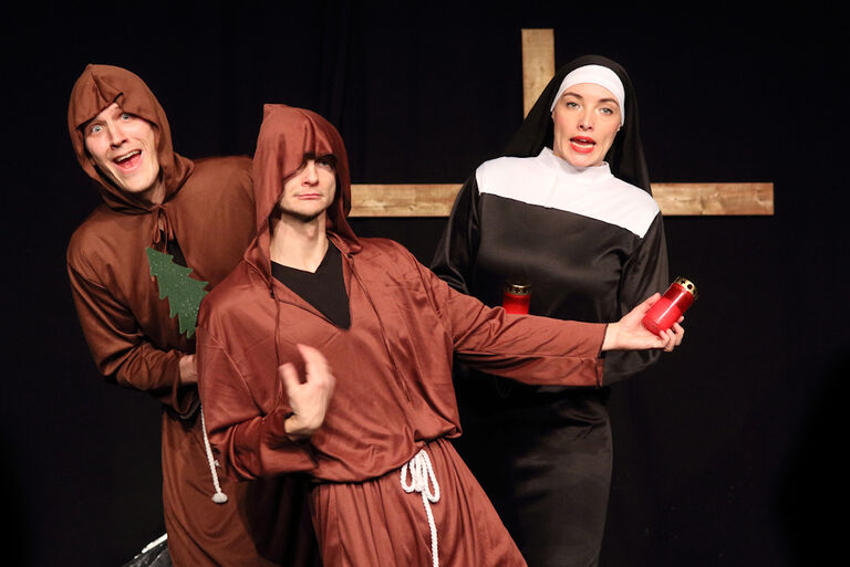 Zwei Schauspieler als Mönche und eine Schauspielerin als Nonne verkleidet stehen in seltsamen Posen vor einem Holzkreuz