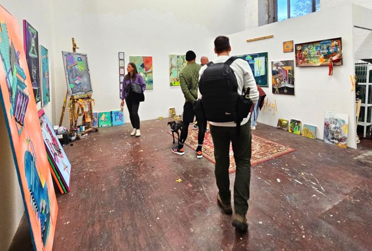 In einem Atelier hängen viele abstrakte Gemälde oder sind an die Wände gelehnt und werden von Besucherinnen und Besuchern betrachtet.