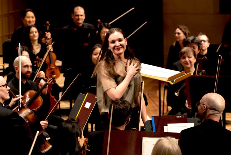 Foto des Orchesters im Konzertsaal, in dessen Mitte eine junge Frau aufgestanden ist und glücklich lächelt