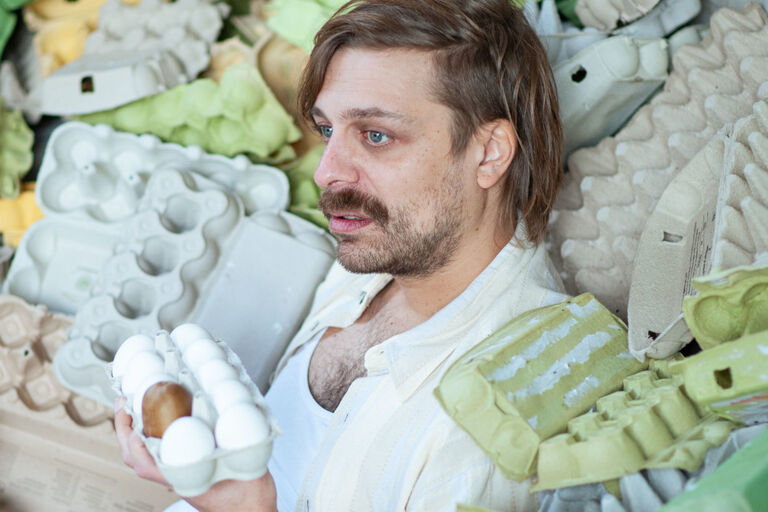 Ein Mann, der zwischen vielen leeren Eierkartons sitzt und einen vollen in der Hand hält, schaut mit ängstlichem Blick in die Ferne.