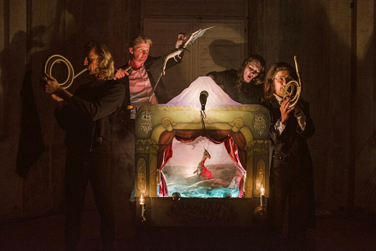 Um eine kleine Puppenbühne stehen vier Schauspielerinnen und Schauspieler, spielen Instrumente und bewegen Puppen