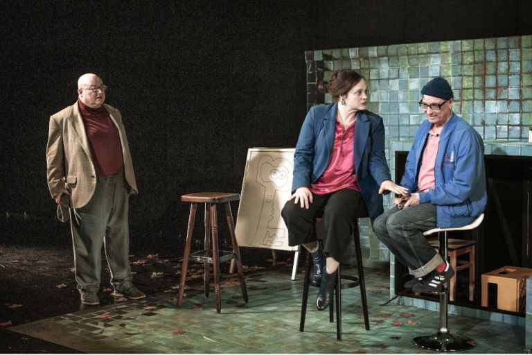 Zwei Schauspieler und eine Schauspielerin stehen oder sitzen vor einer gekachelten Wand