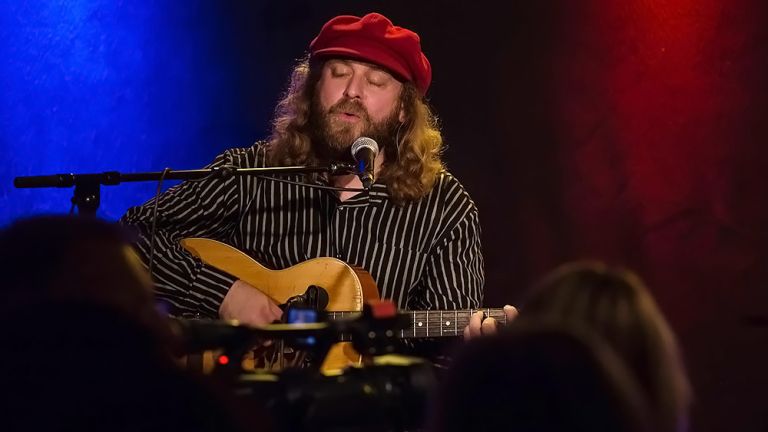 Der Musiker sitzt bei einem Konzert mit roter Schiebermütze und geschlossenen Augen auf der Bühne und spielt Gitarre.