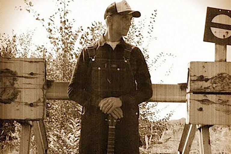 Der Musiker steht in Arbeitskleidung und mit einer Gitarre vor einem Rammbock am Ende eines Bahngleises.