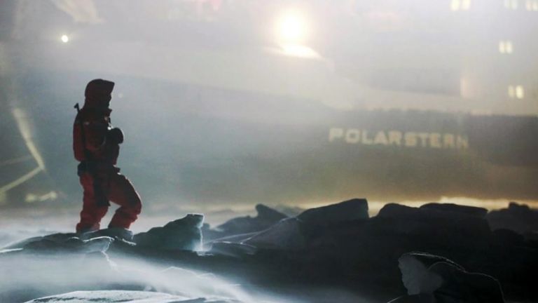 Im Vordergrund ist eine Person im roten Overall mit umgehängten Gewehr, dahinter groß das hell erleuchtete Forschungsschiff "Polarstern" zu sehen.