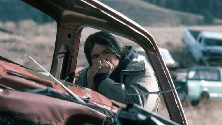 Filmszene, in der sich ein Angehöriger der Cheyenne auf ein Autowrack lehnt und Mundharmonika spielt.