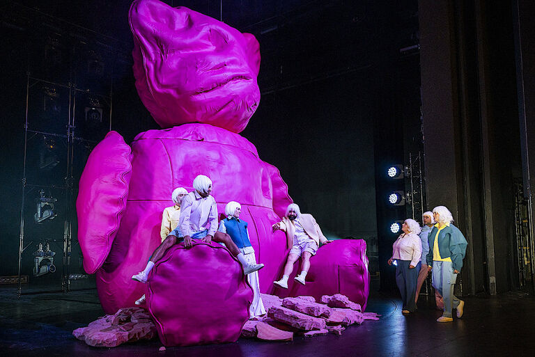 Mehrere Schauspielerinnen und Schauspieler sitzen auf oder stehen an einem überdimensionalen, pinkfarbenen Teddybär.