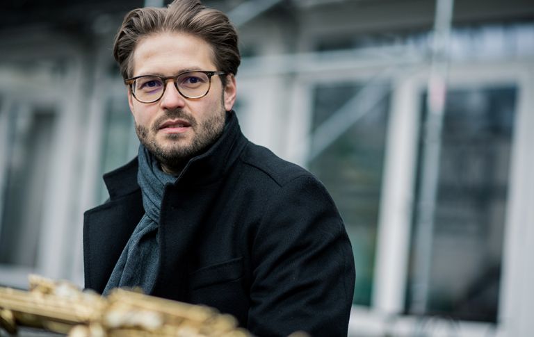 Der Musiker sitzt im Wintermantel im Freien und hat auf einem Tisch vor sich sein Saxophon zu liegen.