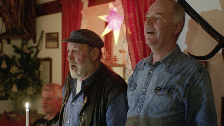 In einer Filmszene singen drei ältere Männer gemeinsam in einer weihnachtlich geschmückten Stube.