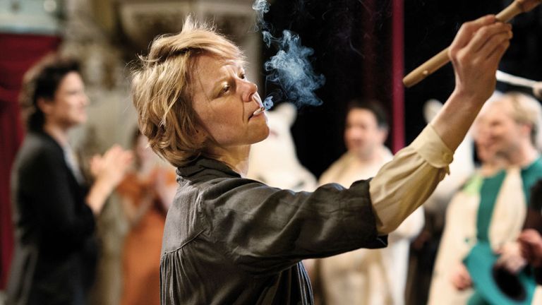 Die Protagonistin steht vor ein paar Menschen mit brennender Zigarette im Mund an einer Staffelei und malt.