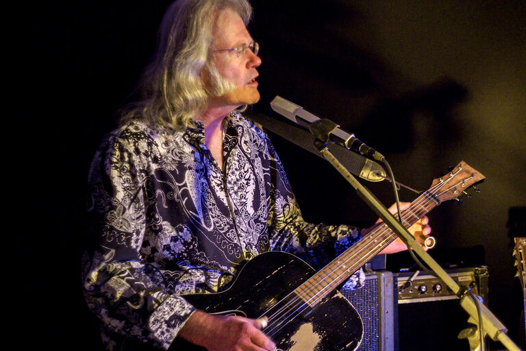 Der Musiker steht bei einem Auftrit mit seiner Gitarre zwischen mehreren Boxen und Instrumenten.