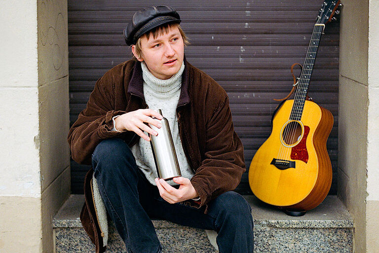 Der Sänger sitzt mit einer Thermoskanne und der Gitarre neben sich auf der Stufe eine geschlossenen Ladentür.