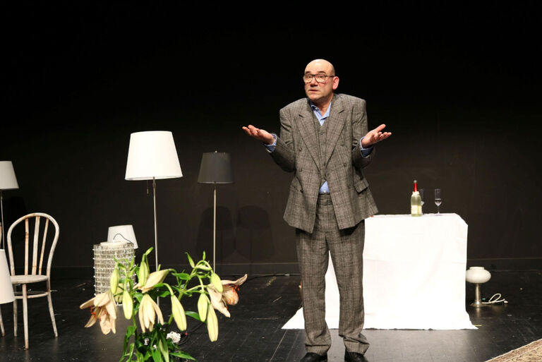 Ein älterer Mann mit Glatze und Brille steht mit ratlos erhobenen Händen auf der Bühne.