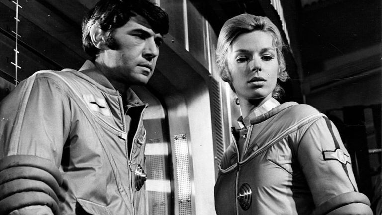 Ein Kosmonaut und eine Kosmonautin in Raumanzgen schauen gebannt auf einen nicht sichtbaren Punkt im Raum.