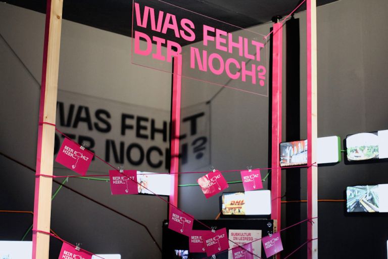 In einem Ausstellungsraum hängen an gespannten Fäden mit Karten und Antworten von Besuchern auf die Frage: Was fehlt dir noch?