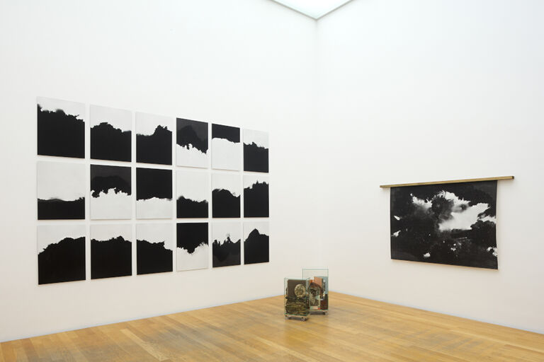 Blick in einen Ausstellungsraum mit Schwarz-Weiß-Fotografien an den Wänden und Gegenständen in Glasbehältern
