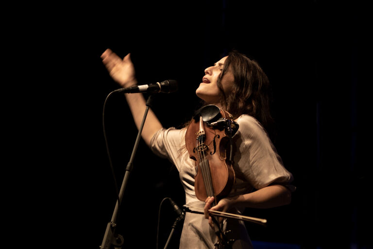 Die Musikerin singt bei einem Auftritt und hat dabei eine Geige im Arm.