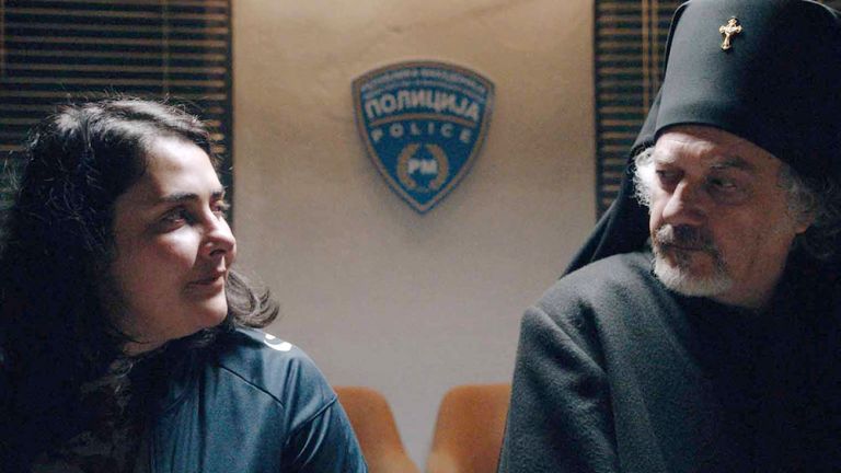 Ein Polizistin und ein Priester im traditionellen schwarzen Gewand sitzen nebeneinander und schauen sich mit ernsten Gesichtern an.