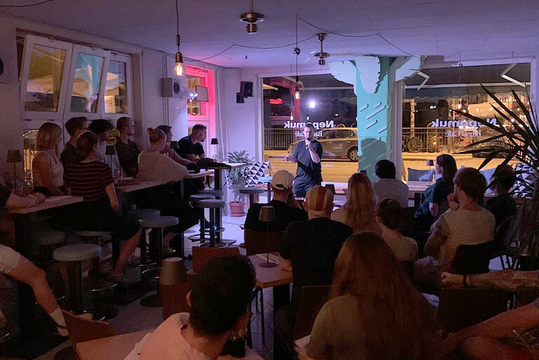 In einer Bar sitzen viele Personen an Tischen und verfolgen die Show eines Comedian.