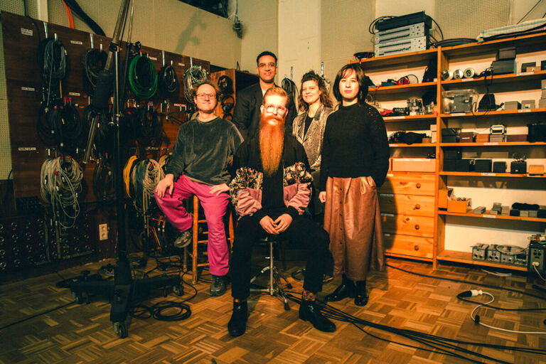 Foto der fünf Musikerinnen und Musiker in einem Studio, an dessen Wänden viele Kabel hängen und Regale mit technischen Geräten stehen