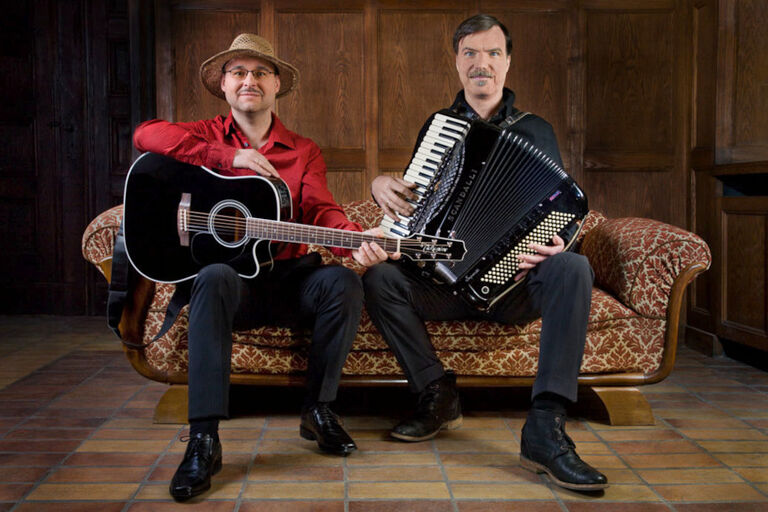 Die beiden Musiker sitzen mit Gitarre und Akkordeon nebeneinander auf dem Sofa.