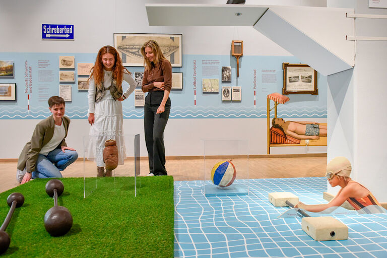 Besucherinnen sehen sich in einer Ausstellung eine nachgestellte Badeszene mit Wiese und Schwimmbecken an.