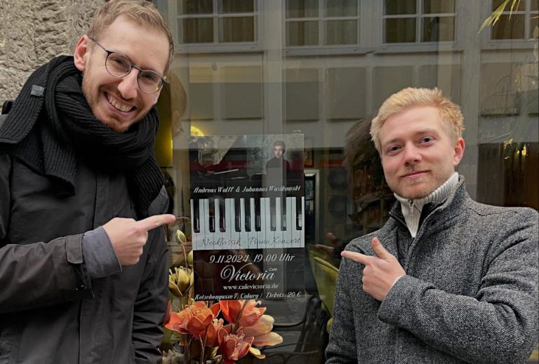Die beiden Pianisten zeigen beide lächelnd auf ein Plakat, das ein gemeinsames Konzert ankündigt.
