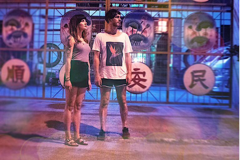 Das Duo steht vor einem Gitter, an dem asiatische Schreiftzeichen angebracht sind.