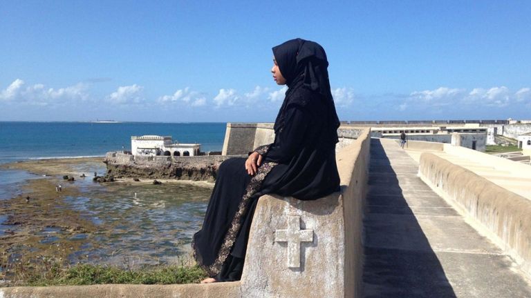 Eine junge Frau in schwarzem Gewand und mit schwarzem Kopftuch sitzt auf einer Mauer am Meer.