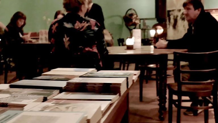 Hinter einem Büchertisch sitzen an weiteren Tischen mehrere Personen in einer Bar.