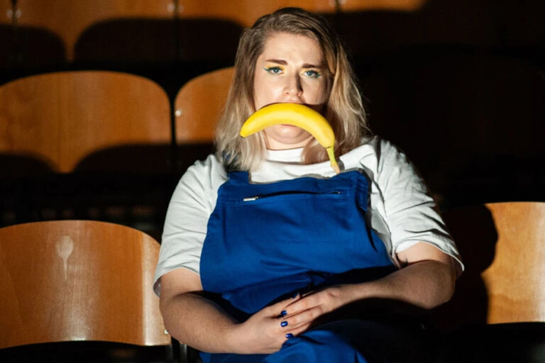 Eine Frau in blauer Latzhose sitzt in im Zuschauersaal eines theaters und hat eine ungeschälte Banane quer im Mund.