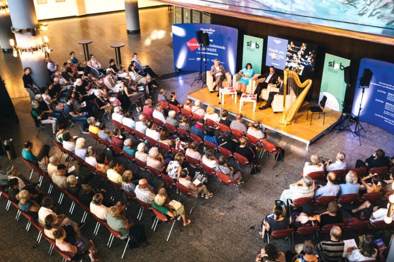 Im Foyer des Gewandhauses sitzen Besucherinnen und Besucher in Stulreihen und verfolgen das Gespräch auf einen Podium.