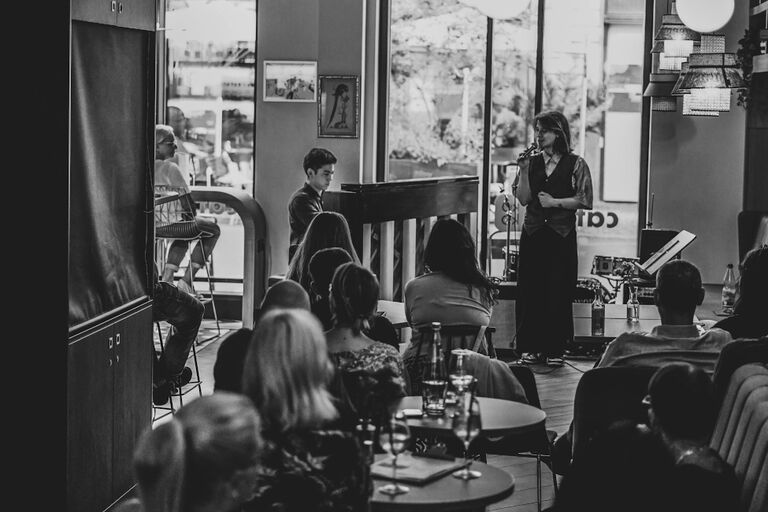 In einem Café sitzen die Gäste an Tischen und verfolgen das Konzert eines Pianisten und einer Sängerin.