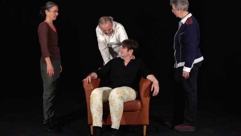 Vier Schauspieler*innen auf einer dunklen Bühne, von denen eine in der Mitte in einem Sessel sitzt und die anderen um sie herumstehen.