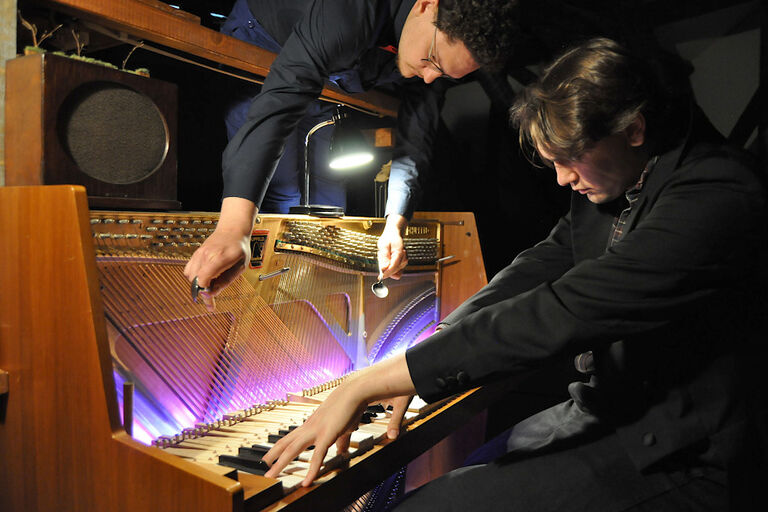 Zwei Musiker an einem Klavier, von denen einer auf den Tasten spielt und der andere mit Löffeln auf den Saite im Inneren des Instrumentsn