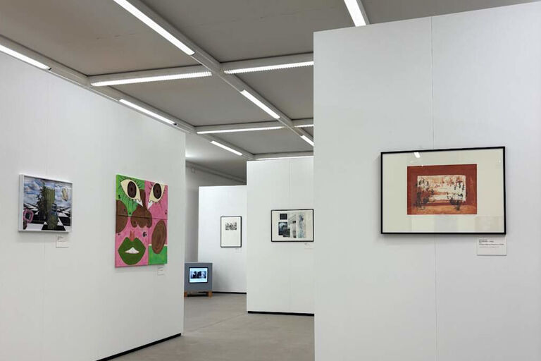 In einem Ausstellungsraum hängen Gemälde und Grafiken an weißen Wänden. In einer Ecke sind auch Bilder auf einem Monitor zu sehen.