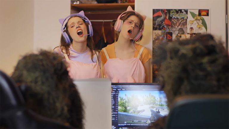 Zwei junge Frauen in pastellfarbener Kleidung singen vor zwei Personen, die an einem Computer sitzen.