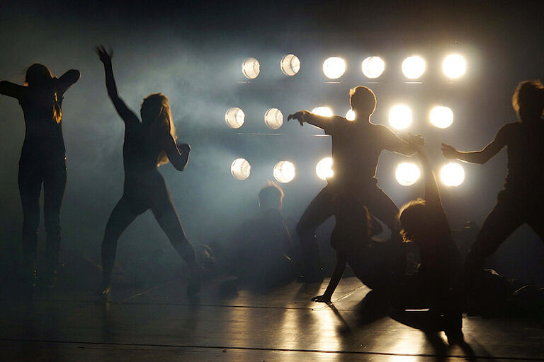 Im Gegenlich von Scheinwerfern sieht man die Umrisse von tanzenden Schauspielerinnen und Schauspielern