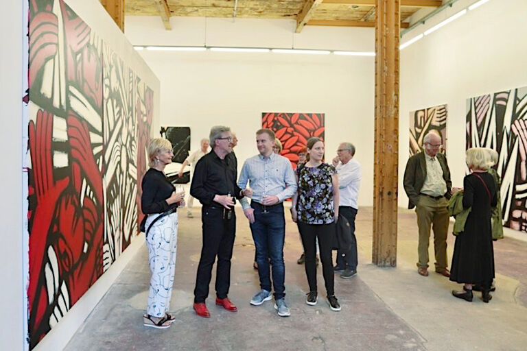 Foto mit dem Maler der zur Eröffnung seiner Ausstellung mit Besucherinnen und Besuchern spricht.