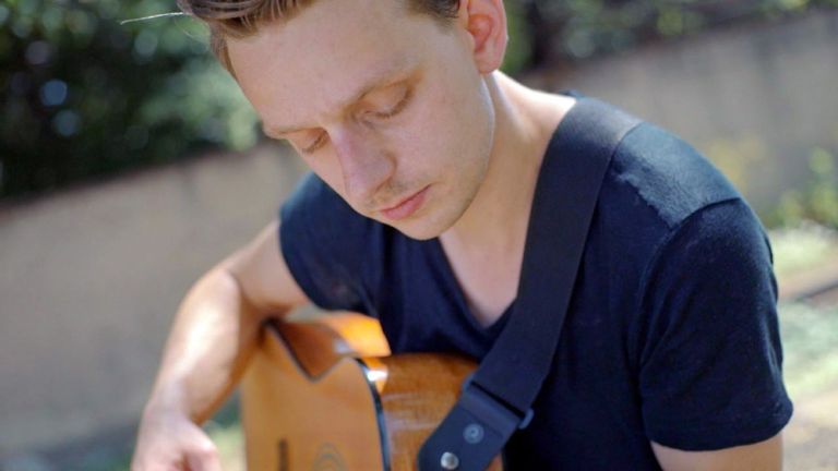 Der Musiker sitzt im T-Shirt mit seiner Gitarre an einem Wasserlauf.